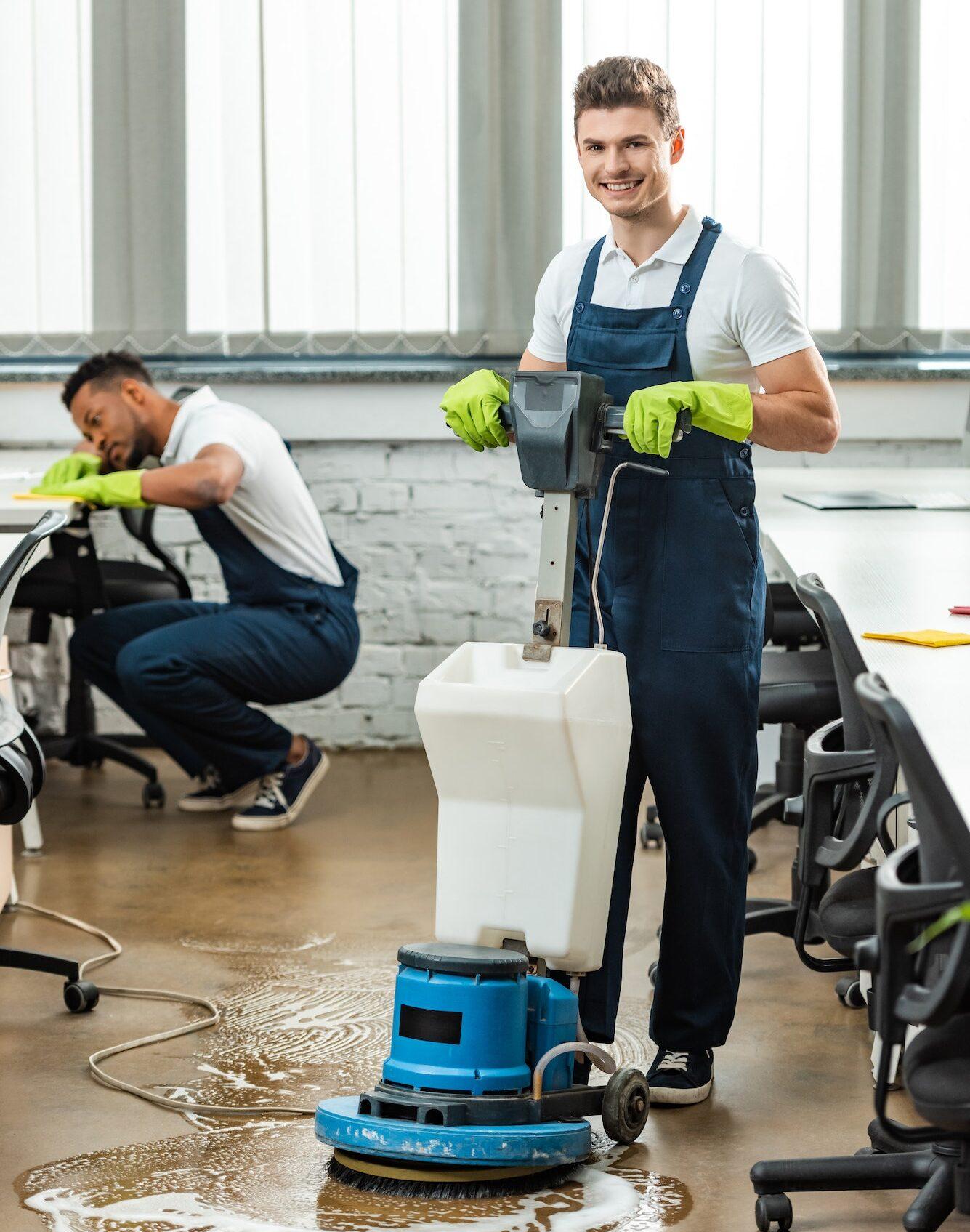 happy cleaner washing floor with cleaning machine while colleague wiping desk